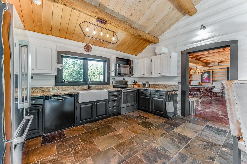 Kitchen with black and white cabinets, granite countertops, a large stainless steel fridge, and a window above the sink with a view of trees outside. There is a doorway leading to a living area with wooden floors and walls.