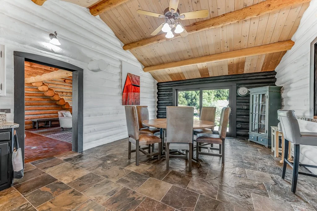 Dining room with stone tile floor, wooden-beamed ceiling, and log walls. A round wooden table with six chairs, a large window, and a blue display cabinet are visible.