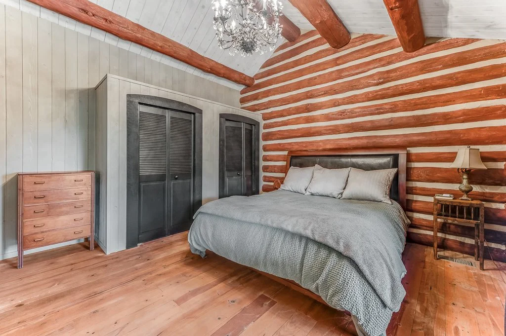 A rustic bedroom with a wooden log wall behind the bed, a chandelier hanging from a sloped ceiling, a gray bedspread with pillows, a small wooden nightstand with a lamp, a wooden dresser, and a closet with louvered doors.