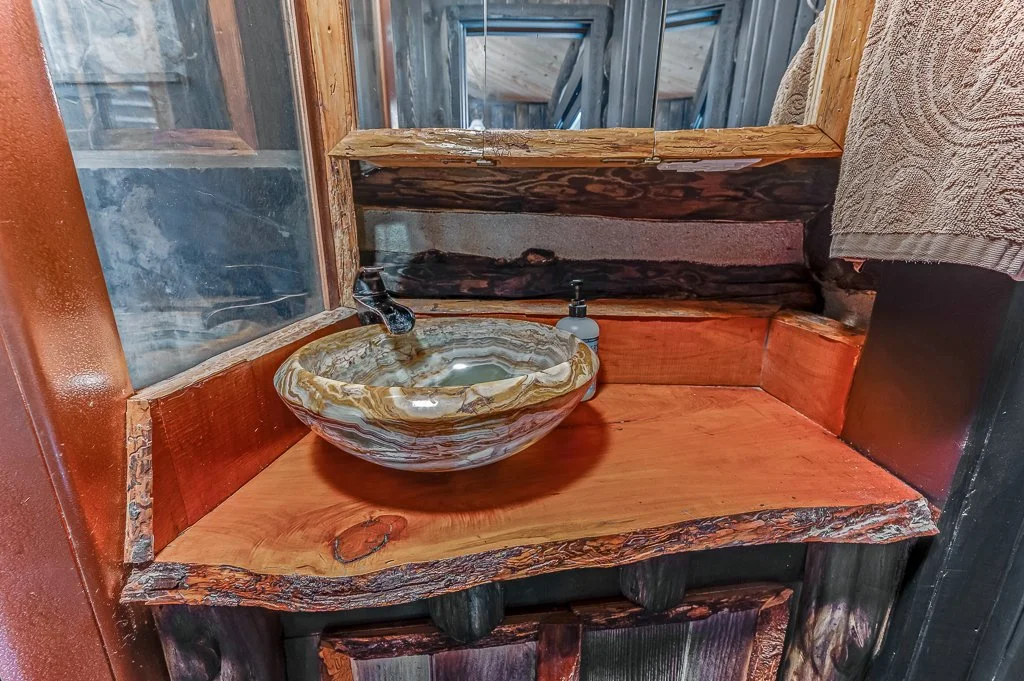 A rustic wooden sink area with a oval stone sink, a soap dispenser, and a small faucet, surrounded by wooden walls and a mirror.
