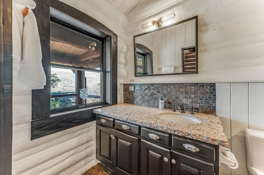 Bathroom with a granite countertop vanity, dark cabinets, a mosaic tile backsplash, a large mirror, a window with black frame, and a towel hanging on the wall.