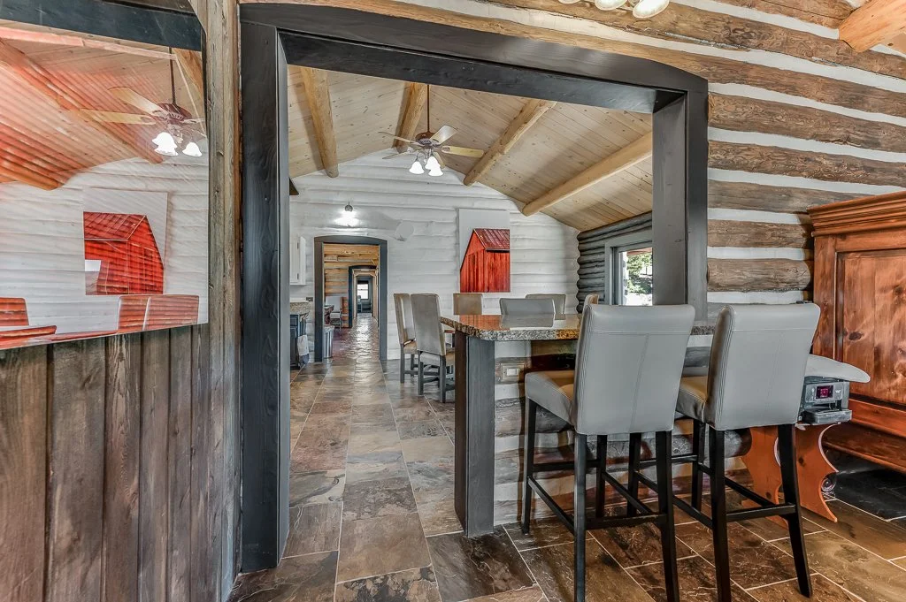 Interior view of a rustic log cabin with a kitchen and dining area, showing wooden logs on the wall, tile flooring, and bar stools at a counter.
