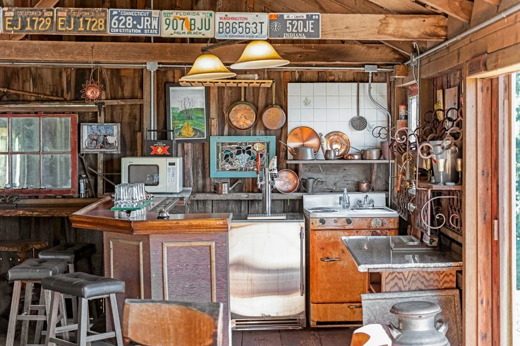 A rustic kitchen with wooden walls and ceiling, decorated with vintage license plates, copper pots, and wall art. Contains a small stove, microwave, and a bar counter with stools.