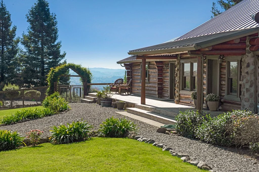 A log cabin with a metal roof, surrounded by a landscaped yard with shrubs, plants, and a gravel pathway, overlooking a scenic view of trees and mountains in the distance.
