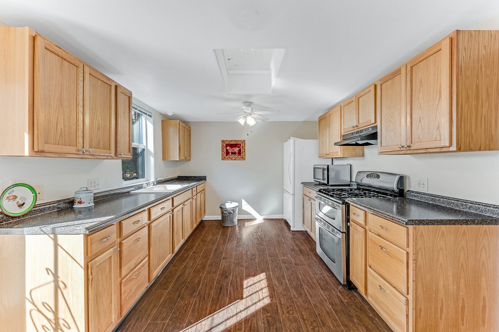 Kitchen with wooden cabinets, black granite countertops, white refrigerator, stainless steel stove, microwave, overhead fan, window, and hardwood flooring.