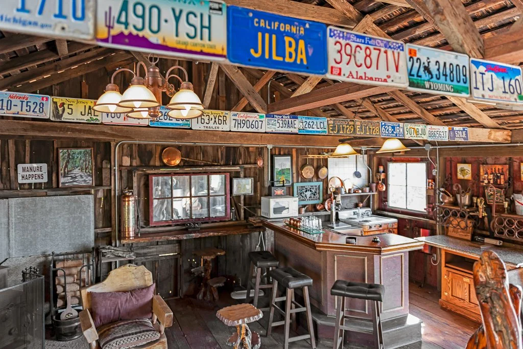 Interior of a rustic bar or man cave with wooden walls and ceiling, decorated with vintage license plates from various states, wooden furniture, bar stools, and eclectic décor including a sign that says 'Manure Happens'.
