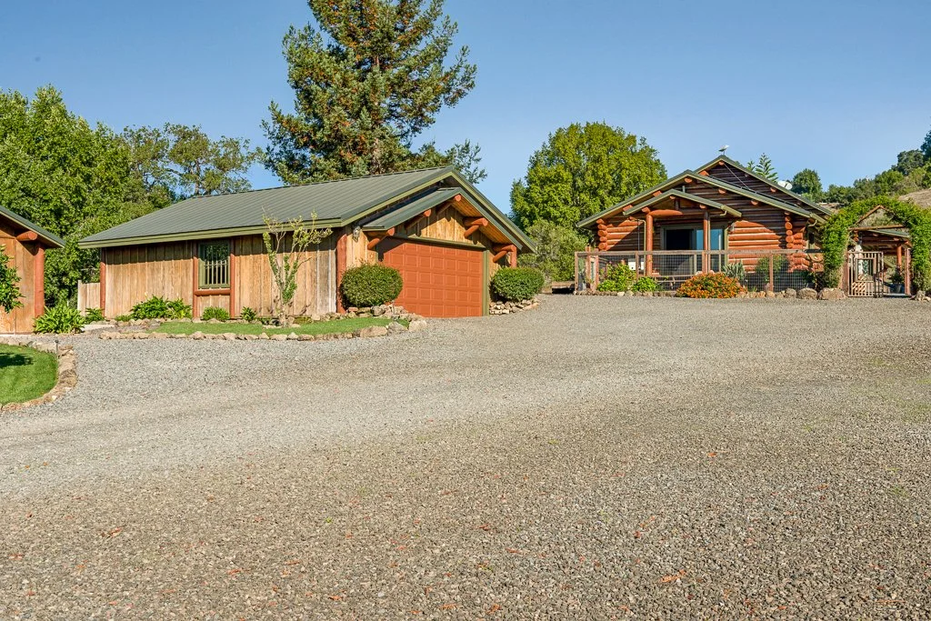 Two houses in a rural setting with a gravel driveway in the foreground and trees in the background.