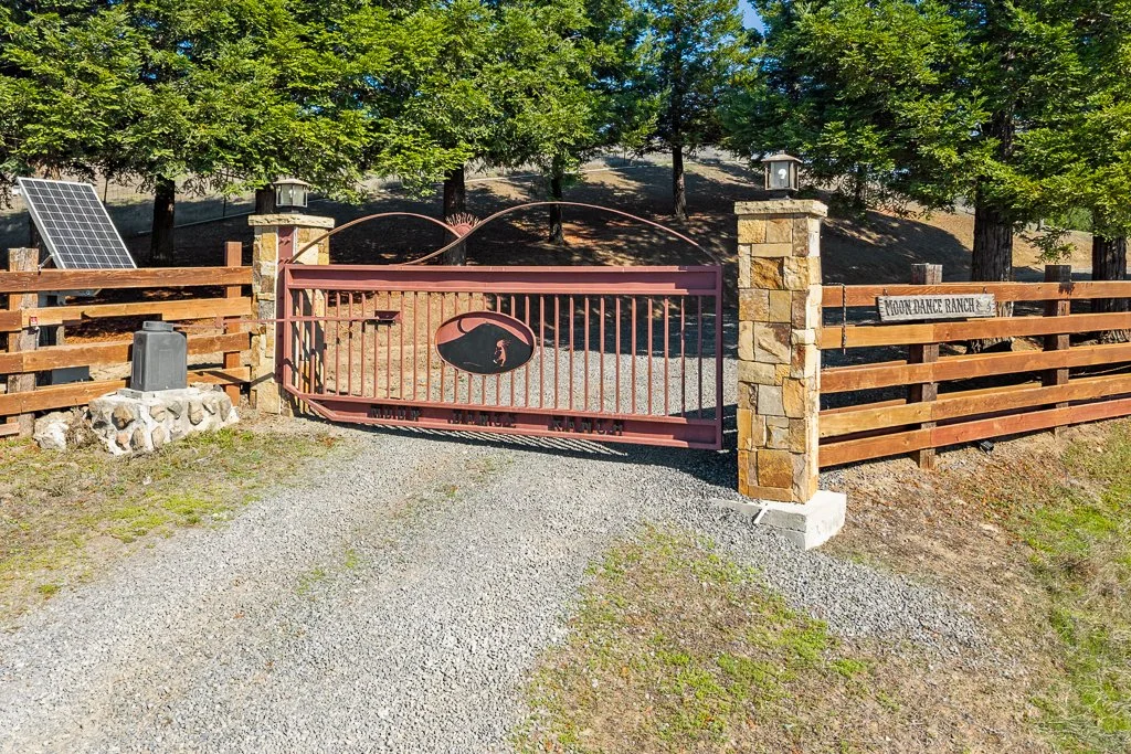 Entrance gate to MoonDance Ranch with a wooden fence, stone pillars, and a sign, surrounded by green trees.