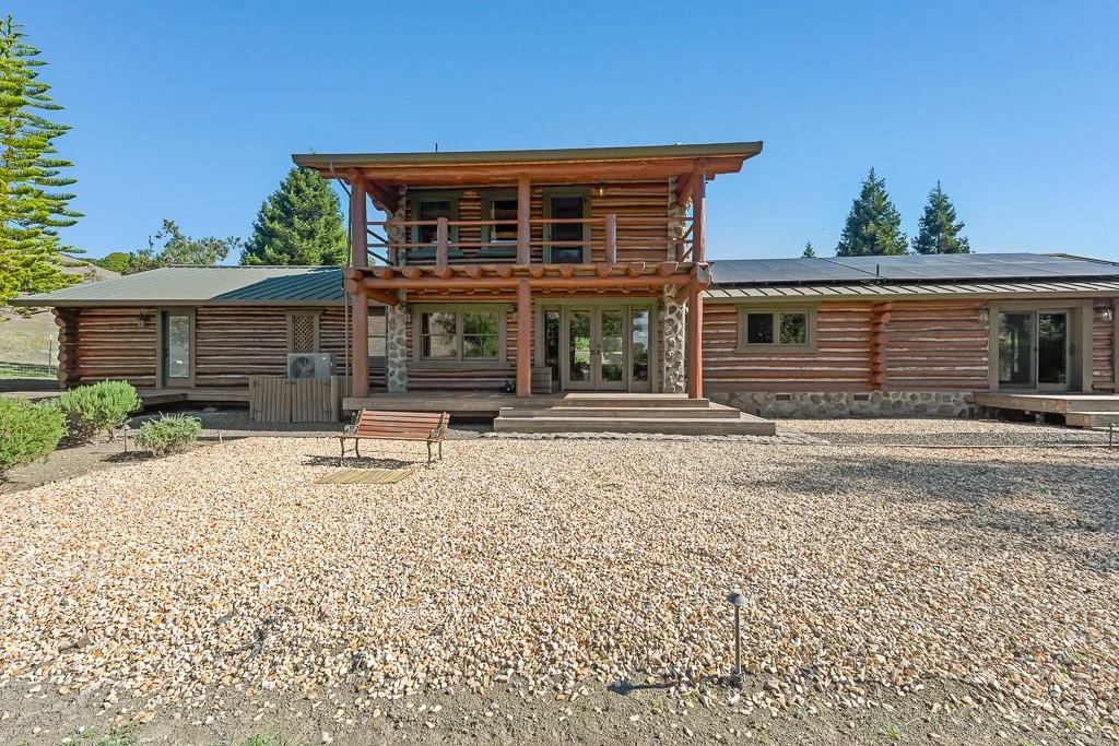 Front view of a two-story wooden log cabin with a balcony, surrounded by gravel ground and some greenery, with a clear blue sky in the background.