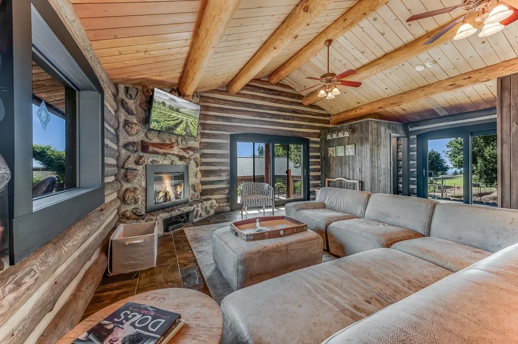 Living room with a large beige sectional sofa, a stone and wood fireplace, a wall-mounted TV, a ceiling fan, large windows with views of greenery, and a wooden ceiling with exposed beams.