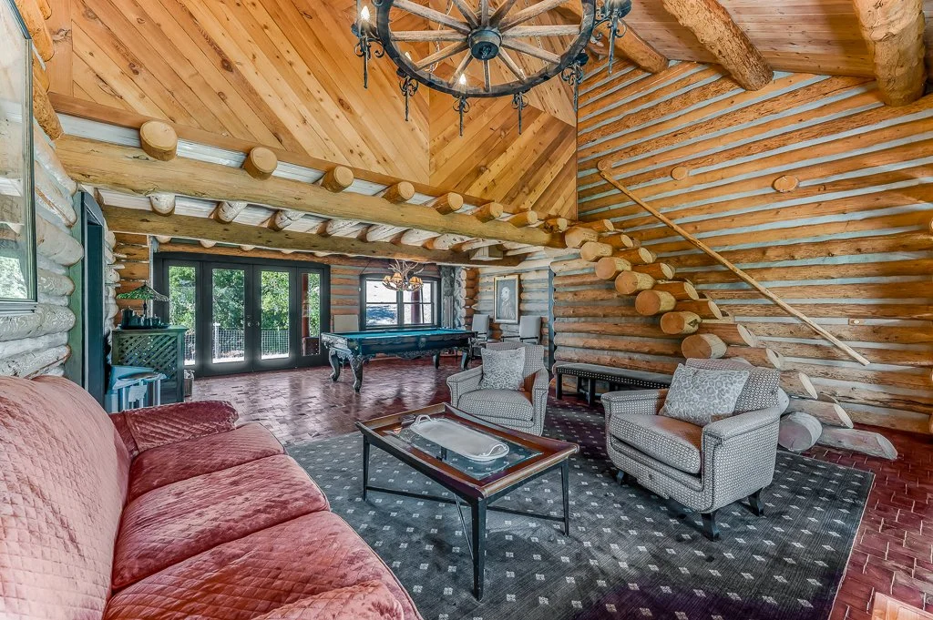 Inside a rustic log cabin with log walls and wooden ceiling beams, featuring a chandelier, a red upholstered sofa, patterned armchairs, a glass coffee table, a black pool table, and large glass doors leading outside.