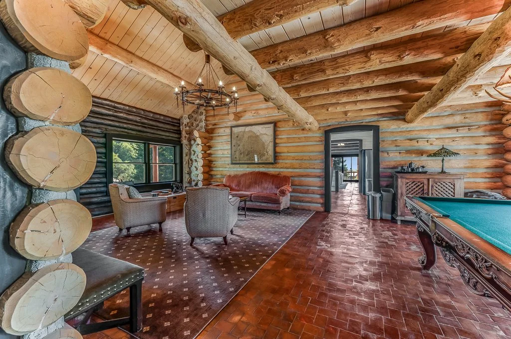 Interior of a rustic log cabin living room with wooden logs on the walls and ceiling, a pool table, a window with views of green trees, and vintage furniture.