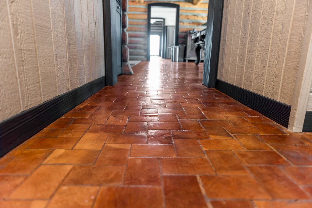 A low-angle view of a hallway with wooden floor tiles and beige walls, leading to a room with furniture and a door in the background.