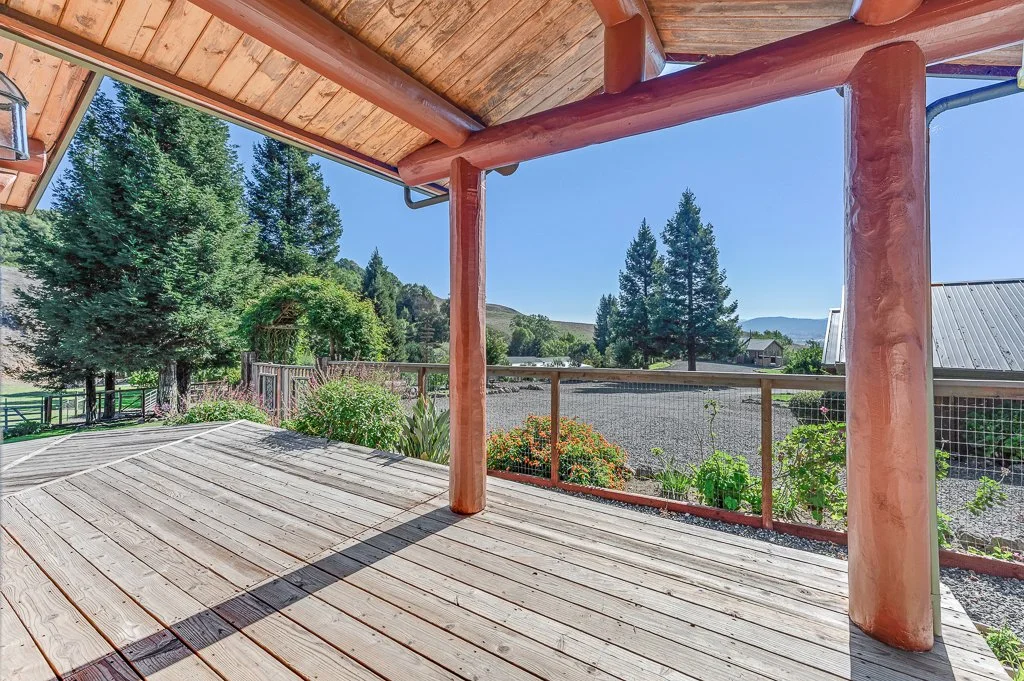 Wooden deck with railing overlooking a rural landscape with trees and hills under a clear blue sky.