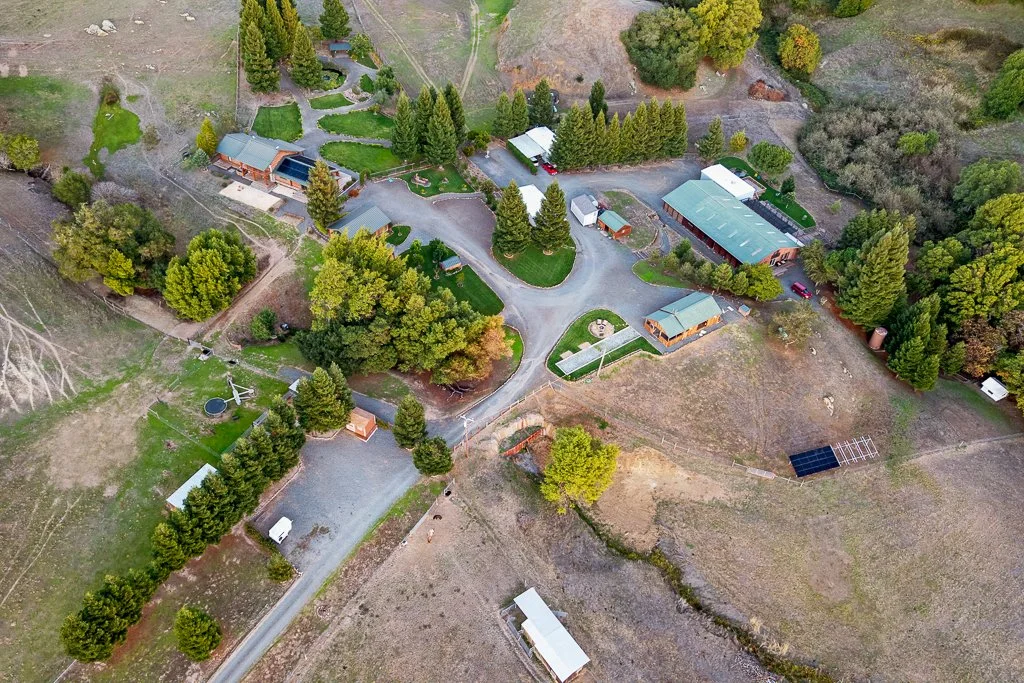 Aerial view of a rural property with multiple buildings, trees, pathways, a trampoline, and solar panels, surrounded by open land and forested areas.