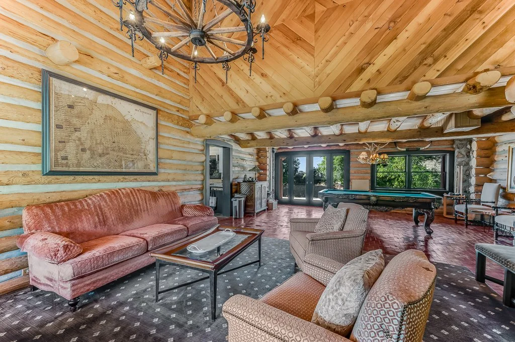 Interior of a log cabin with wooden walls and ceiling, featuring a chandelier, comfortable seating area with a pink velvet sofa and armchairs, a pool table, and large windows with a view of greenery.