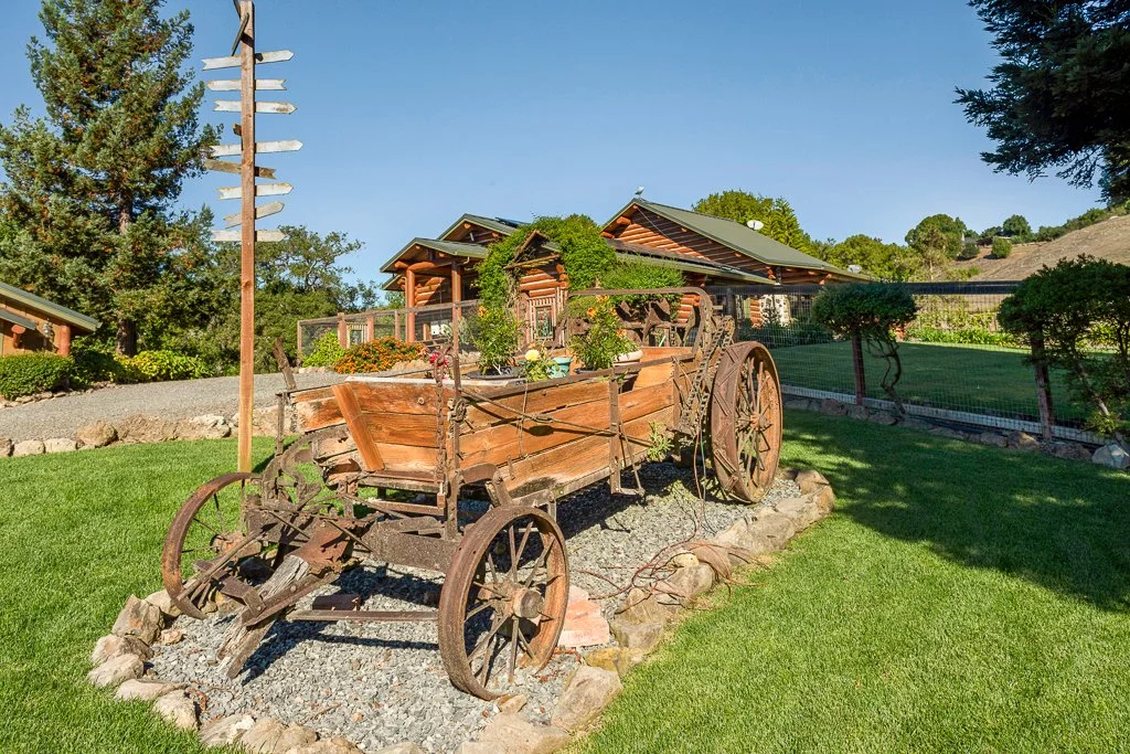 Old wooden wagon with metal wheels used as a garden decoration, placed on a gravel bed surrounded by rocks in a well-maintained yard with a house in the background, trees, and a clear blue sky.