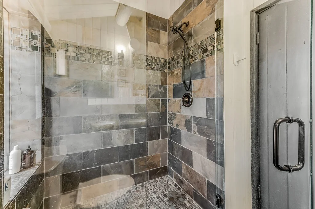 A tiled walk-in shower with a glass door, a showerhead, and a small corner shelf with bottles. The tiles are a mix of gray, beige, and brown shades with a decorative strip of small mosaic tiles near the top.