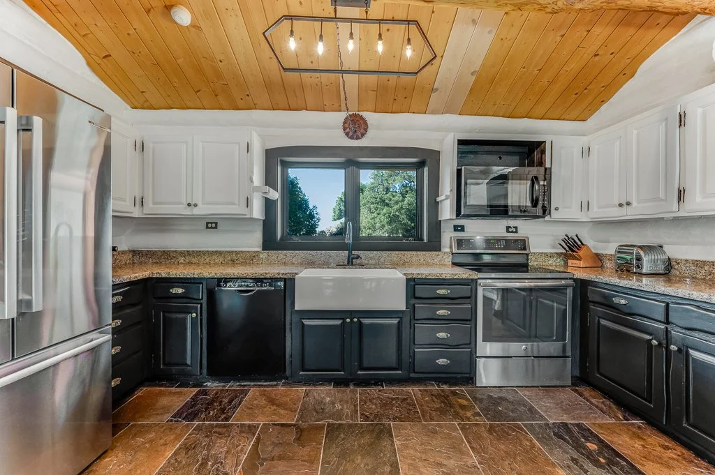 Kitchen with black lower cabinets, white upper cabinets, a granite countertop, a stainless steel refrigerator, a sink under a window, a microwave above the stove, a toaster on the counter, and a sloped wooden ceiling.