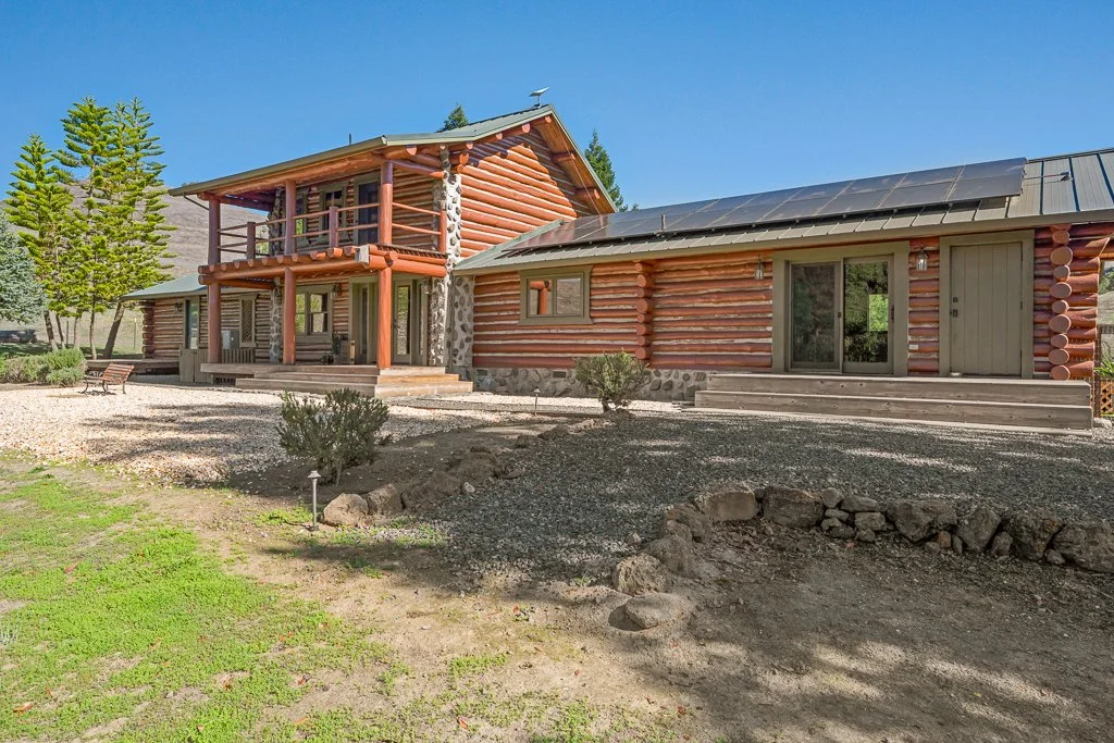 Log cabin-style house with a combination of logs and stone foundation, surrounded by trees and a gravel yard, under a clear blue sky.