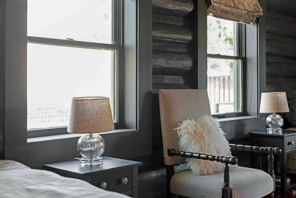 Interior of a rustic bedroom with two windows, beige armchair with a fluffy pillow, and two matching nightstands with glass lamps.