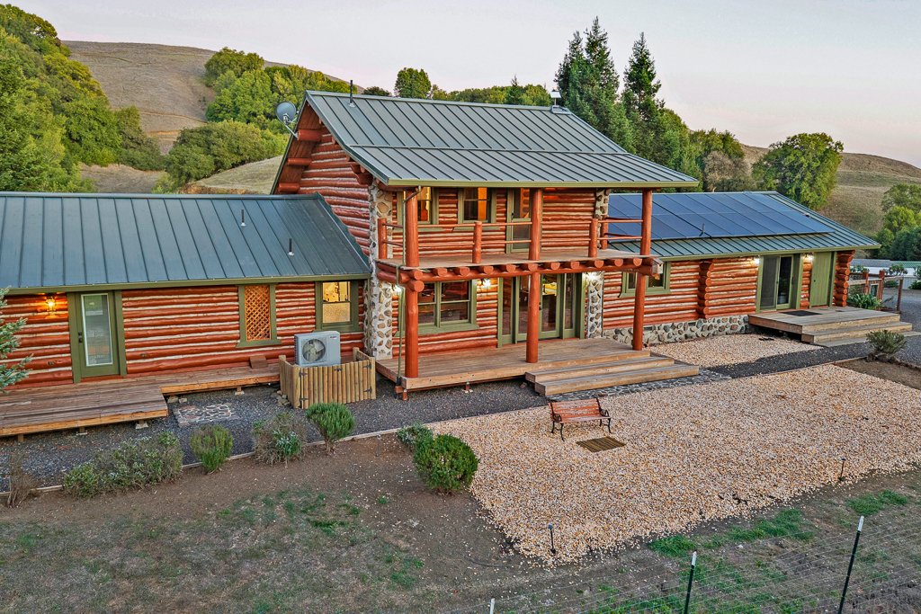 A large log cabin style house with a metal roof, surrounded by a natural landscape of trees and hills, with a gravel yard and wooden deck in the front.