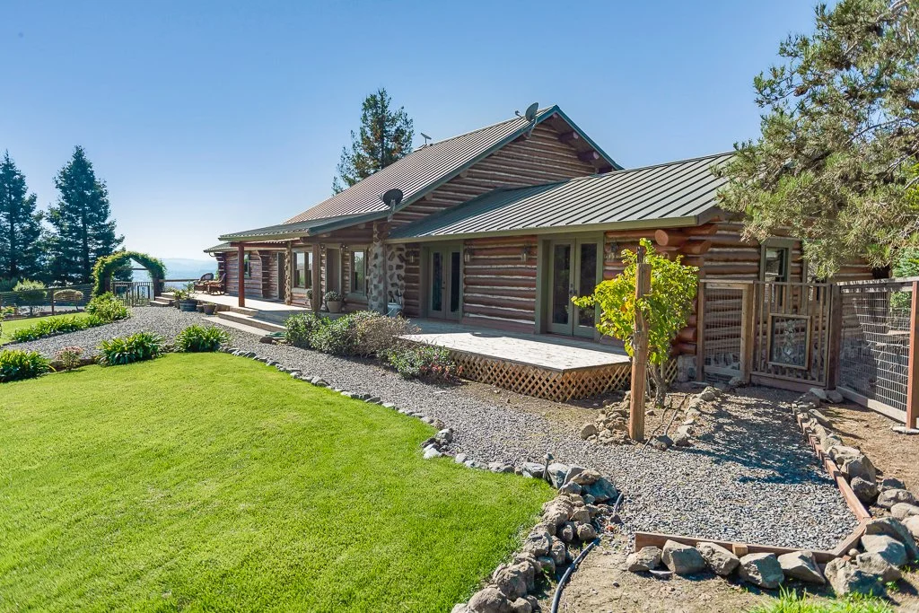 A rustic log cabin with a metal roof and a spacious deck, surrounded by a well-maintained lawn and landscaped garden, with trees and a scenic view in the background under a clear blue sky.