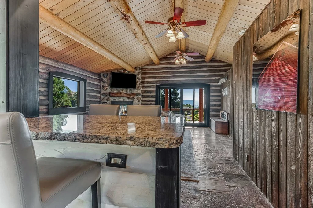 Interior of a rustic log cabin with wooden walls and ceiling, large windows showing greenery and mountains outside, a granite kitchen island, and a sliding door leading to an outdoor patio.