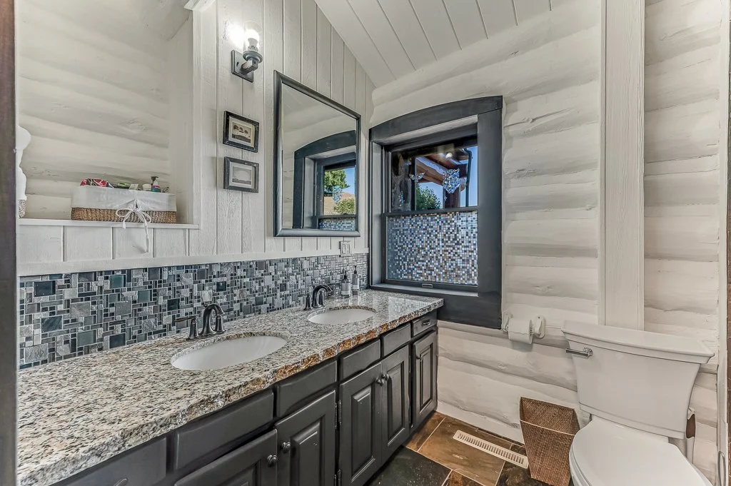 Bathroom with double sinks, granite countertop, mosaic tile backsplash, mirror, window, soap dispenser, wall-mounted light, toilet, and small basket on the floor.