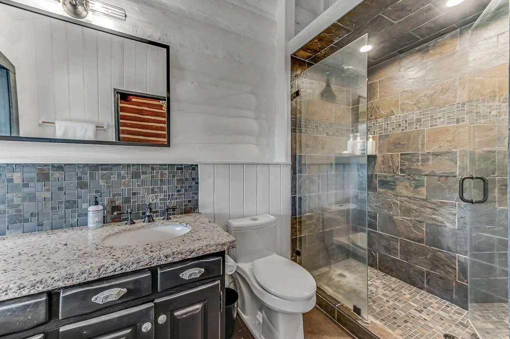 A modern bathroom with a granite countertop sink, a toilet, and a glass-enclosed shower with stone tiles.
