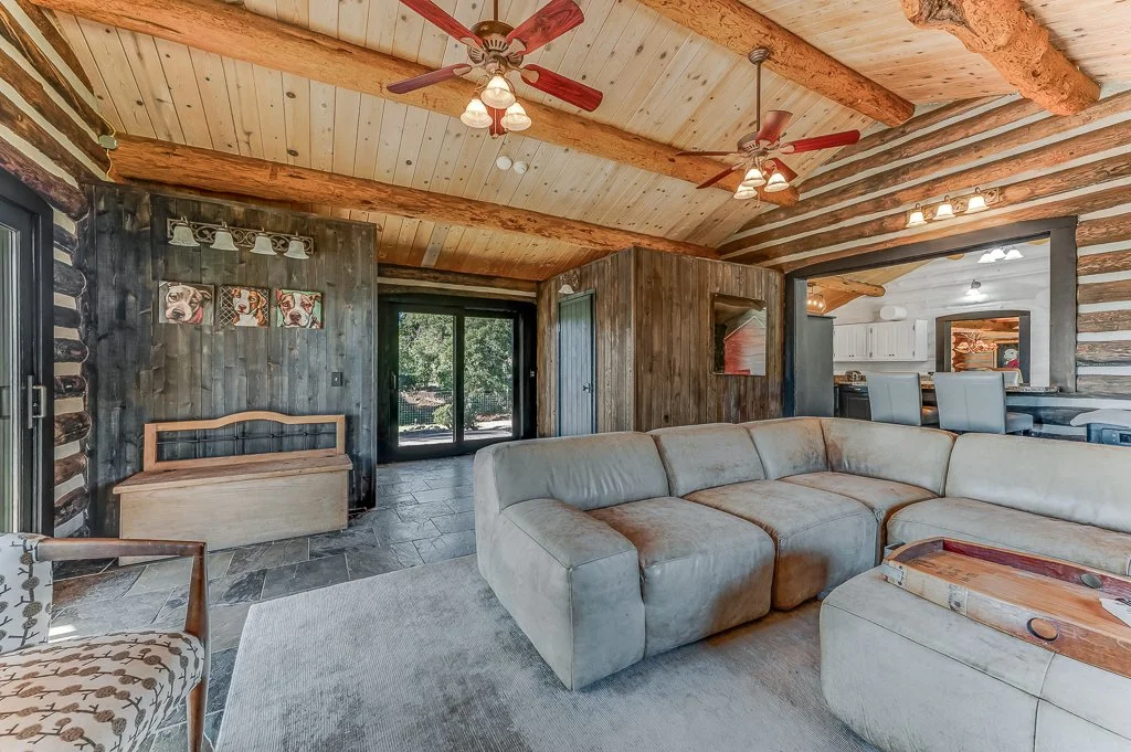 Living room with wood-paneled ceiling, exposed beams, ceiling fans, sectional sofa, and a view of the dining area and kitchen.