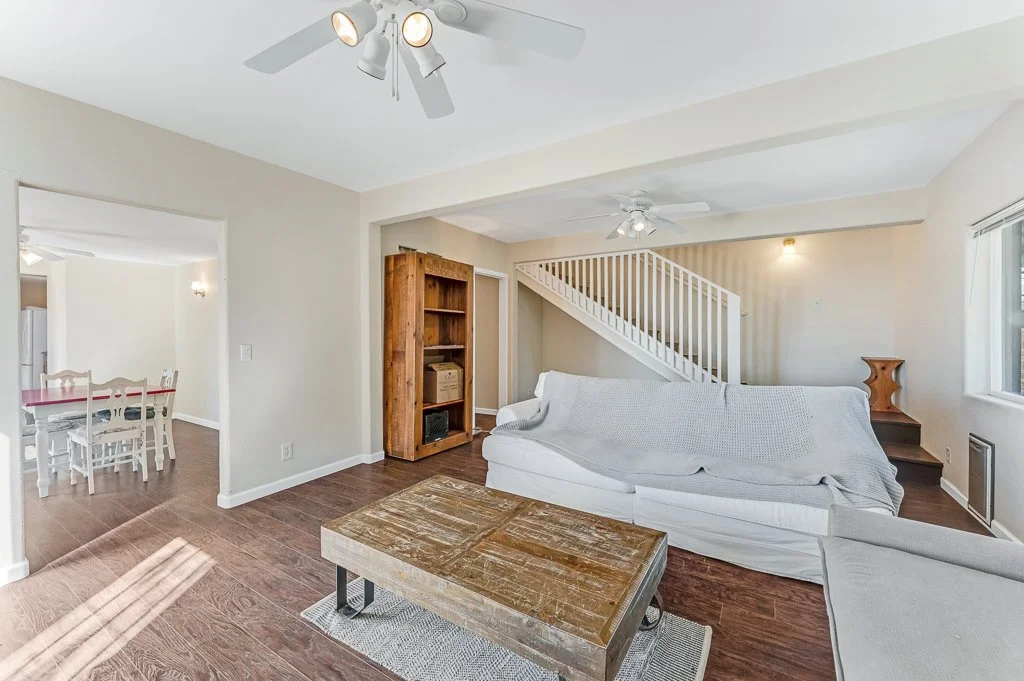 Living room with a white sofa covered with a white slipcover, wooden coffee table, wooden shelving unit, staircase, ceiling fans, a window, and a doorway to a dining room.