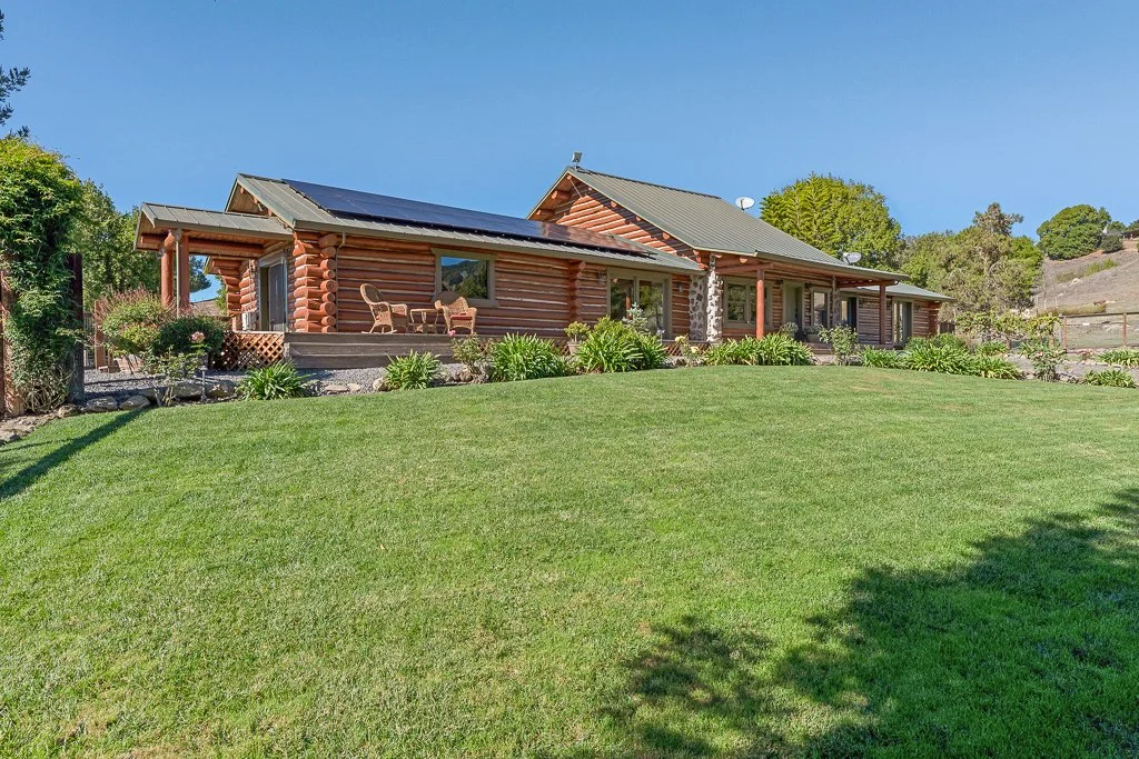 A large Log cabin-style house with solar panels on the roof, surrounded by a well-maintained grassy yard and landscaped garden with bushes and flowers.