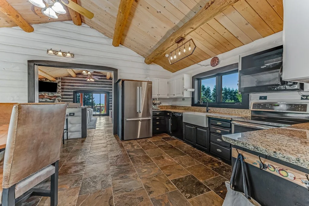 Kitchen with rustic wood ceiling and cabinets, granite countertops, stainless steel refrigerator, black lower cabinets, and a large window with a view of trees.