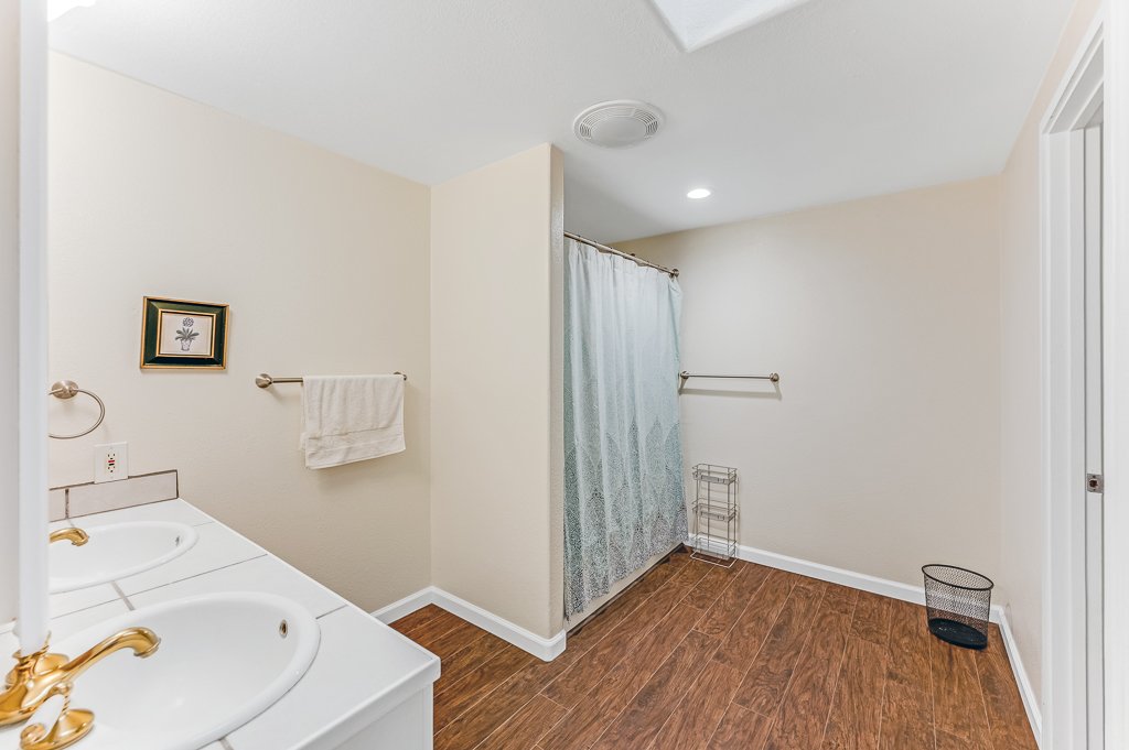 Bathroom with double sinks, a small framed picture on the wall, a towel rack, a shower with a curtain, a small wire rack, and a trash bin on wooden flooring.