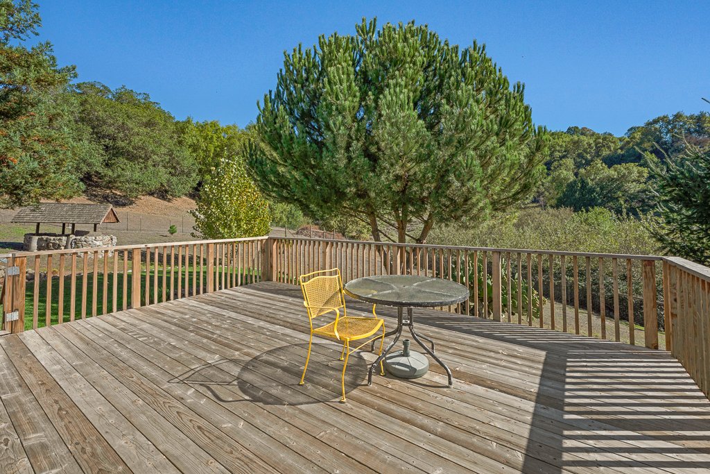 A wooden outdoor deck with a yellow metal chair and a round glass table, overlooking a green landscape with trees and a blue sky.