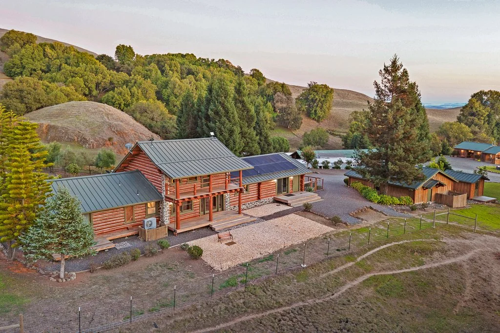 A large log cabin with green metal roofs, situated among trees and hills, with a gravel yard and surrounding fencing.