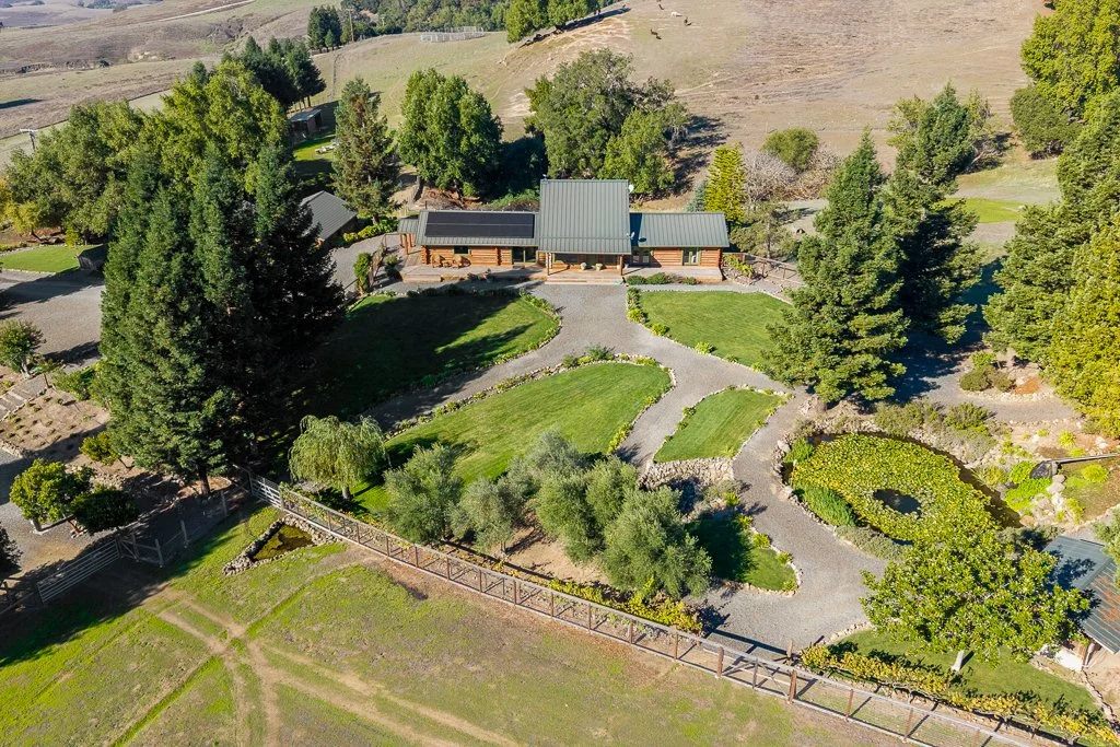 Aerial view of a landscaped garden with curved paths, green grass, trees, and a wooden building in the background, surrounded by countryside.