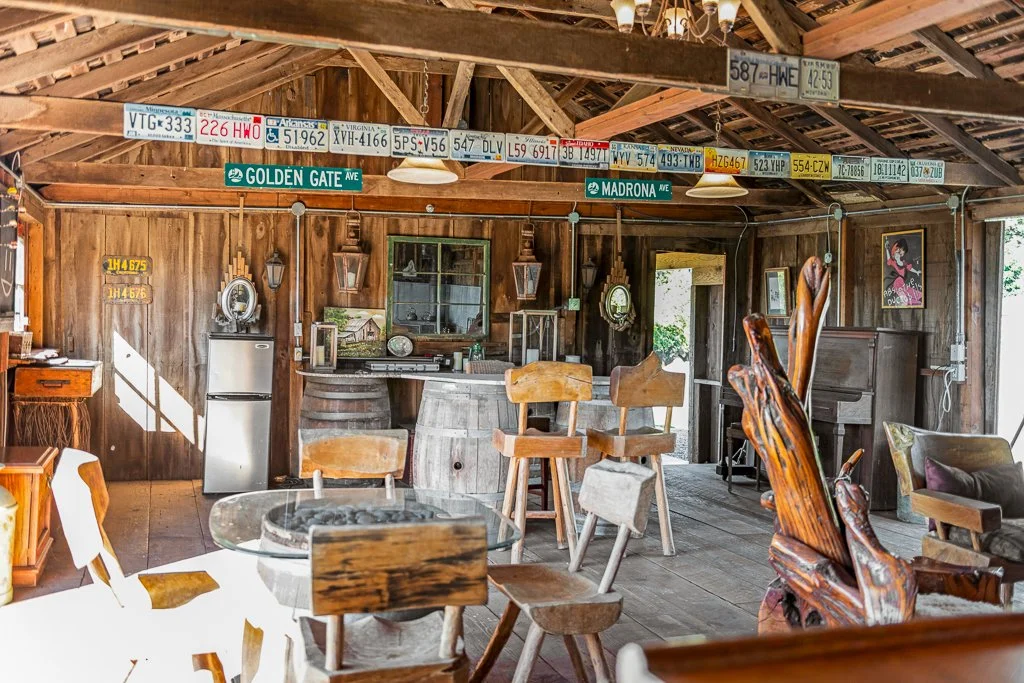 Interior of a rustic wooden cabin with a mix of vintage and handmade furniture, license plates hanging from the ceiling, and various decorative items on the walls and shelves.