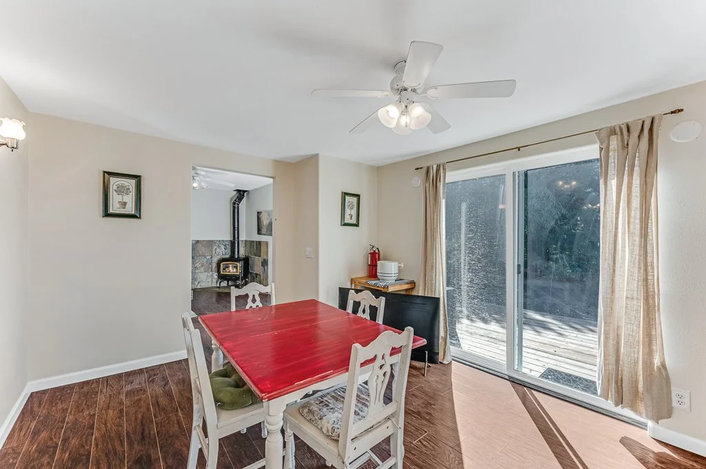Dining room with a red table, white chairs, sliding glass door with curtains, ceiling fan, and a view of a deck outside. A doorway leads to a room with a wood stove.