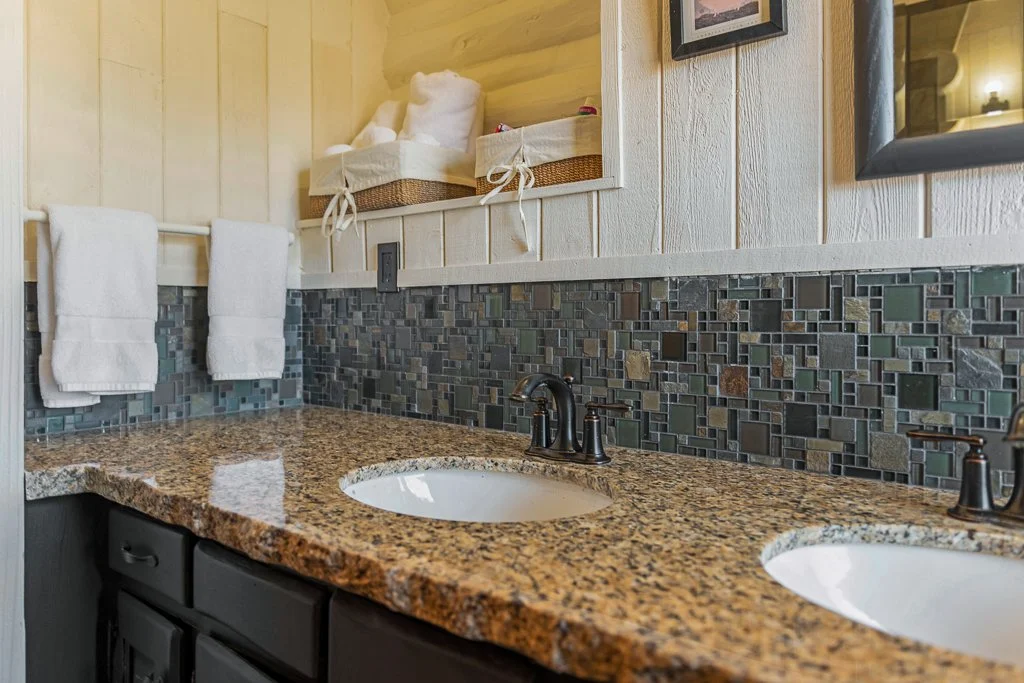 Bathroom vanity with granite countertop, two sinks, brushed bronze faucets, tiled backsplash, two rolled white towels hanging, and baskets with rolled towels and toiletries on shelf above.