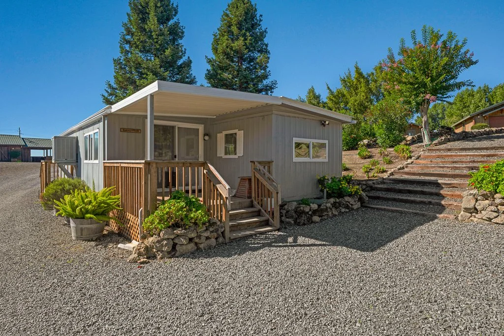 A small, light gray house with a brown wooden porch and railing, surrounded by gravel and greenery, in a sunny outdoor setting with trees and other houses in the background.
