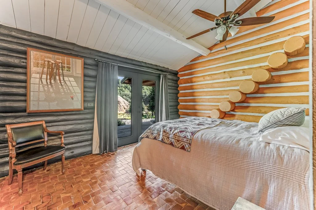 Bedroom with log cabin style walls, a bed with bedding and pillows, a ceiling fan, a framed picture, a chair, and glass double doors with curtains leading outside.