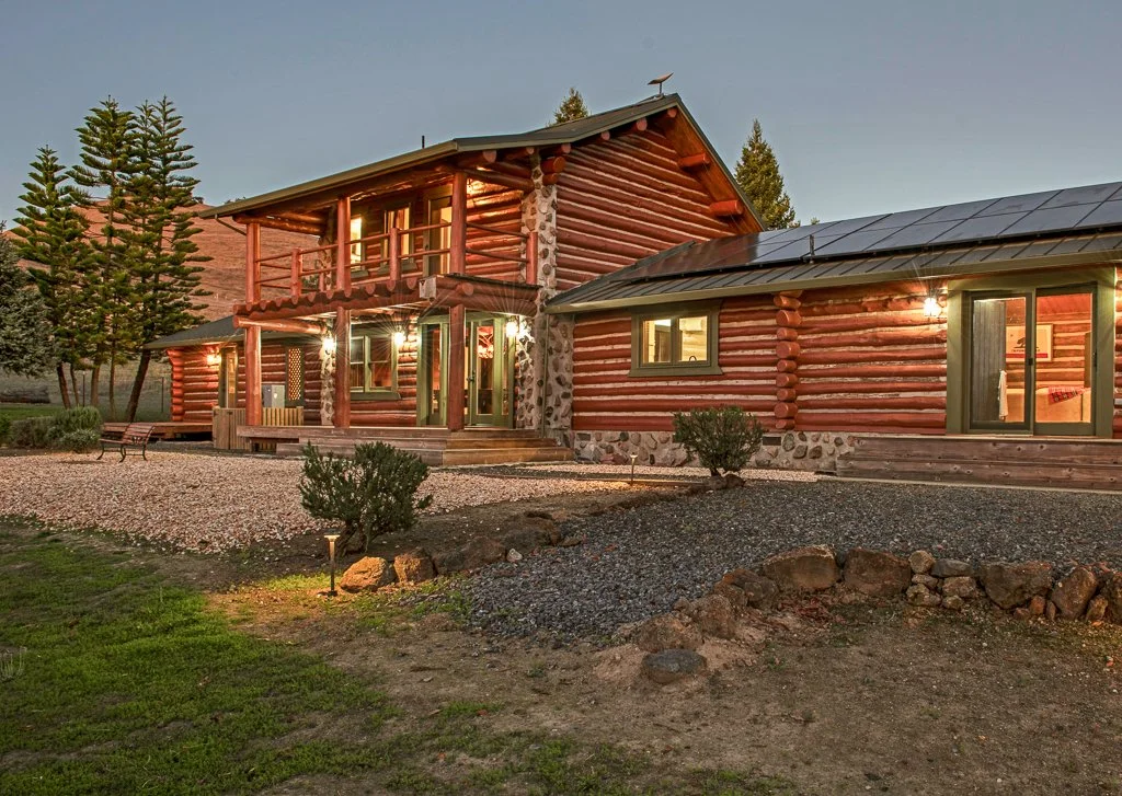 A two-story log cabin with a porch and balcony, surrounded by trees and small bushes, with warm lights illuminating the exterior at dusk.