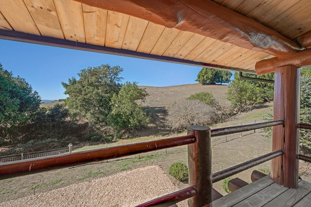A view from a wooden porch with a railing, overlooking a hilly landscape with trees and grassy terrain under a clear blue sky.