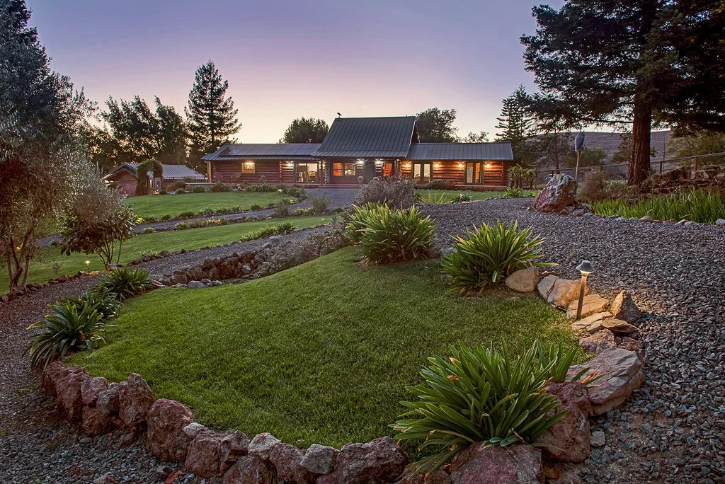 A landscape view of a yard and house during sunset, featuring well-maintained grass, plants, rocks, and a cozy log cabin-style home with lights on.