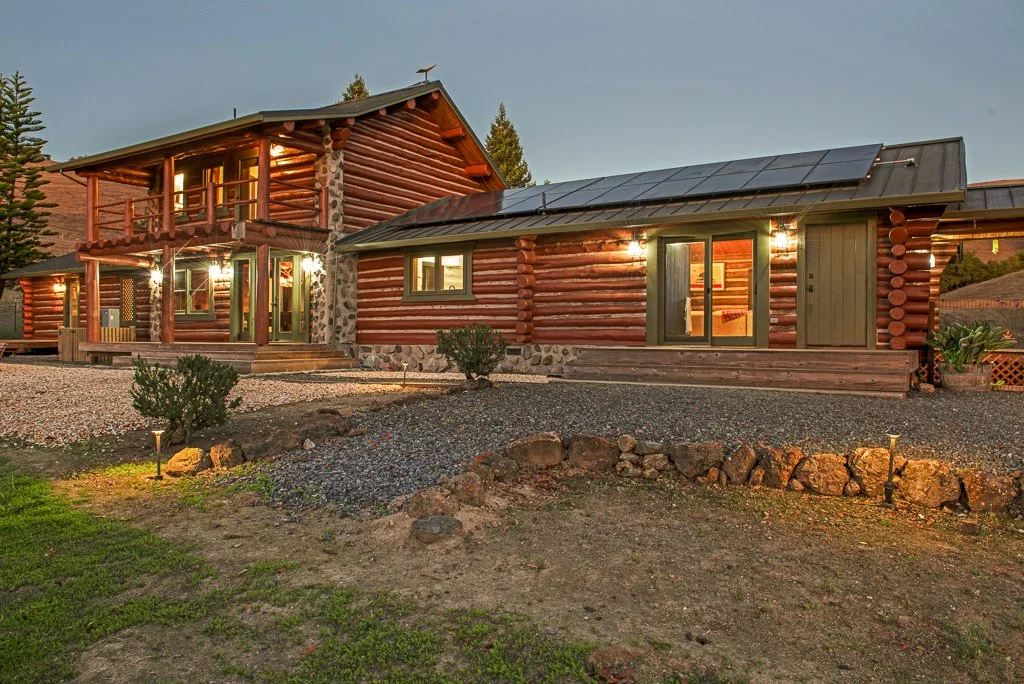 A two-story log cabin house with lit windows, situated in a rural area with trees and mountains in the background during dusk. The house has a balcony, front porch, and solar panels on the roof.