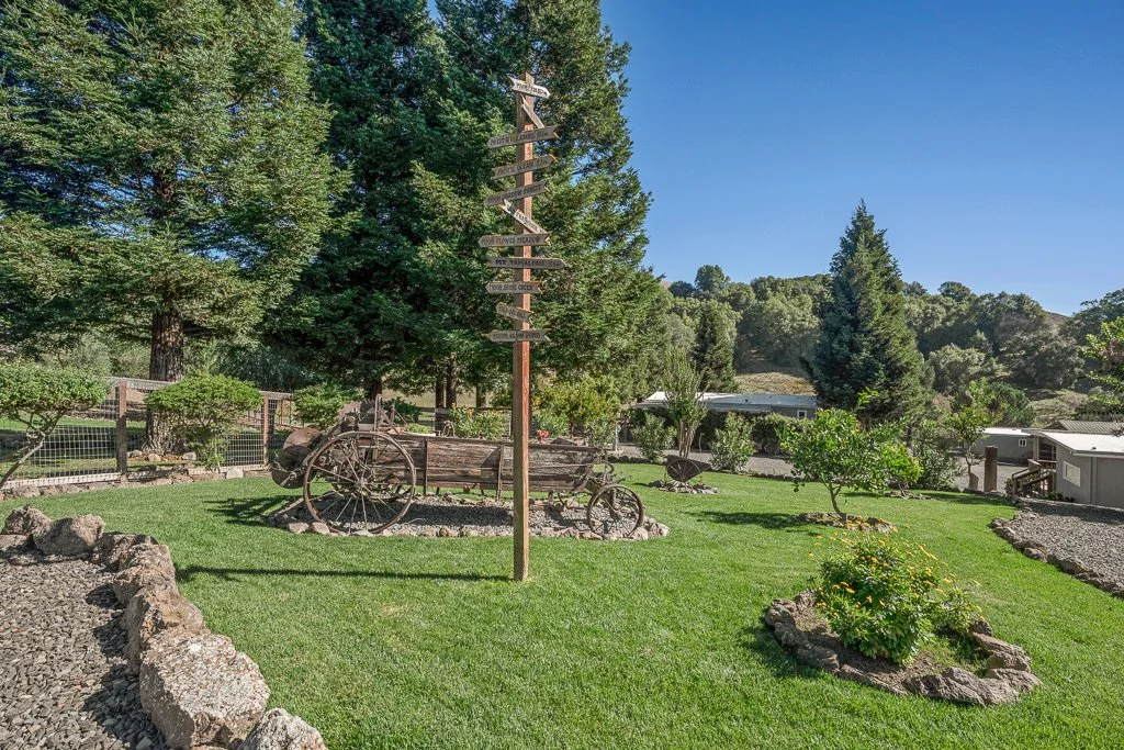 A lush green yard with a tall wooden signpost and vintage wooden farm equipment, including wheels and a cart, surrounded by landscaped plants and trees, with residential buildings and a hill in the background under a clear blue sky.