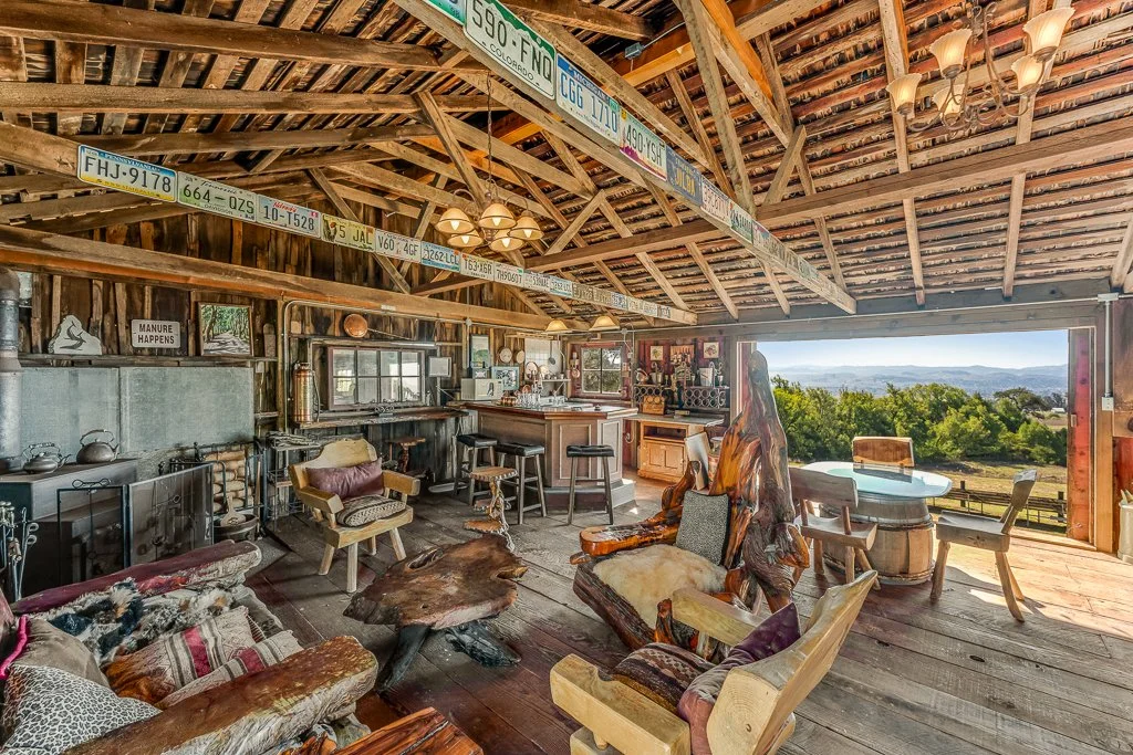 Interior of a rustic wooden cabin with vintage license plates hanging from the ceiling beam, a cozy sitting area with mixed furniture, a bar counter, and large open wall revealing a scenic outdoor landscape with trees and hills.