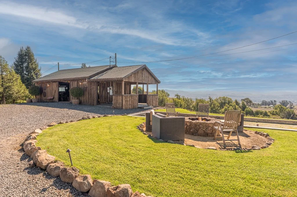A rustic wooden barn with a grey roof, surrounded by a gravel driveway and manicured lawn, with outdoor chairs and a fire pit.